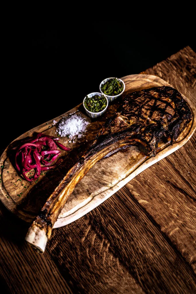 Le Tomahawk, tomahawk steak on a wooden board with chimichurri, red onion and sea salt at Cannibale Royale